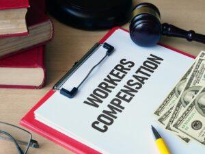 Workers Compensation in black font on white paper in clipboard with money, gavel, and books